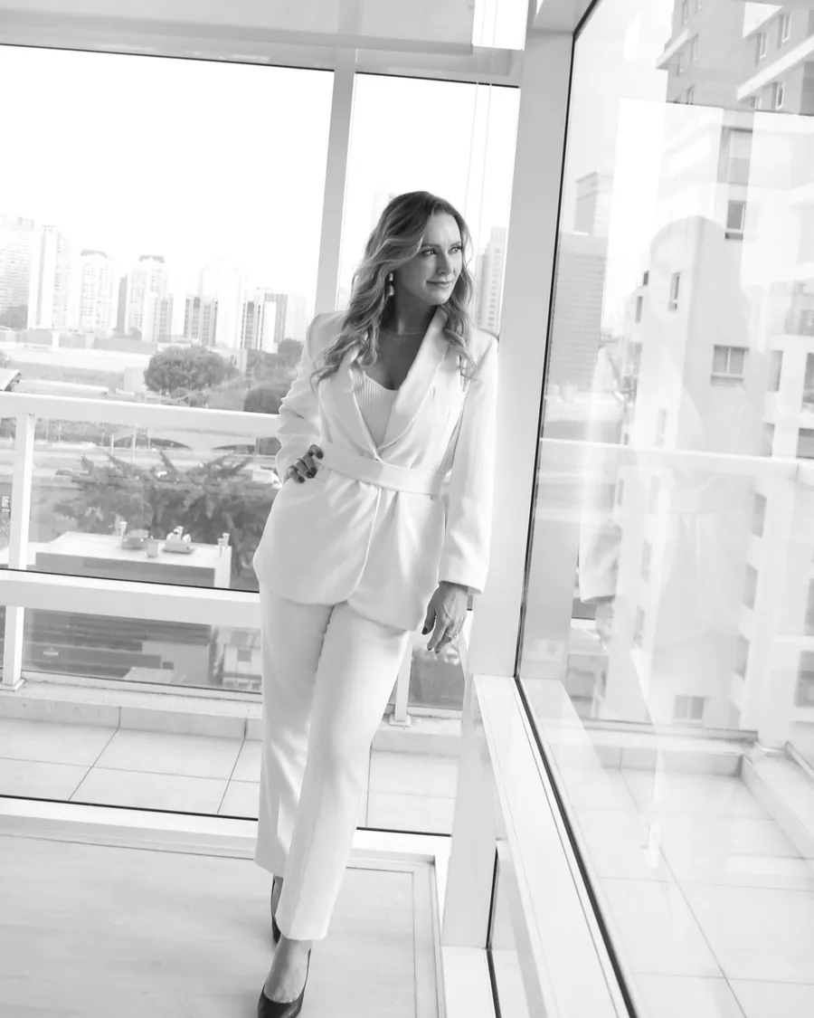 Patrícia Sivieri de terninho branco diante da janela, com a cidade ao fundo